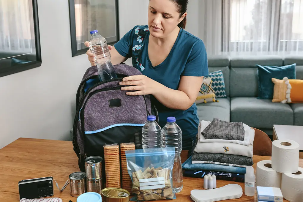 A woman in a blue shirt packs bottled water into a backpack on a table. Surrounding her are canned food, toiletries, folded clothes, and tissues, suggesting preparedness