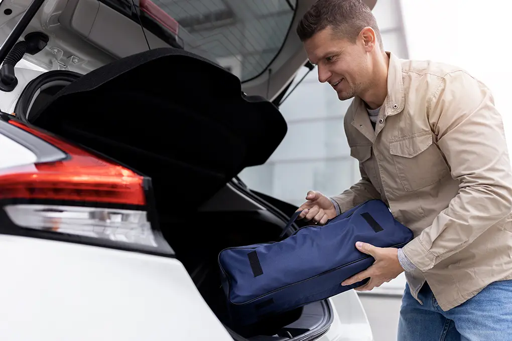 A man in a beige jacket smiles as he places car emergency kit, contained in a blue bag, into the open trunk of a white car