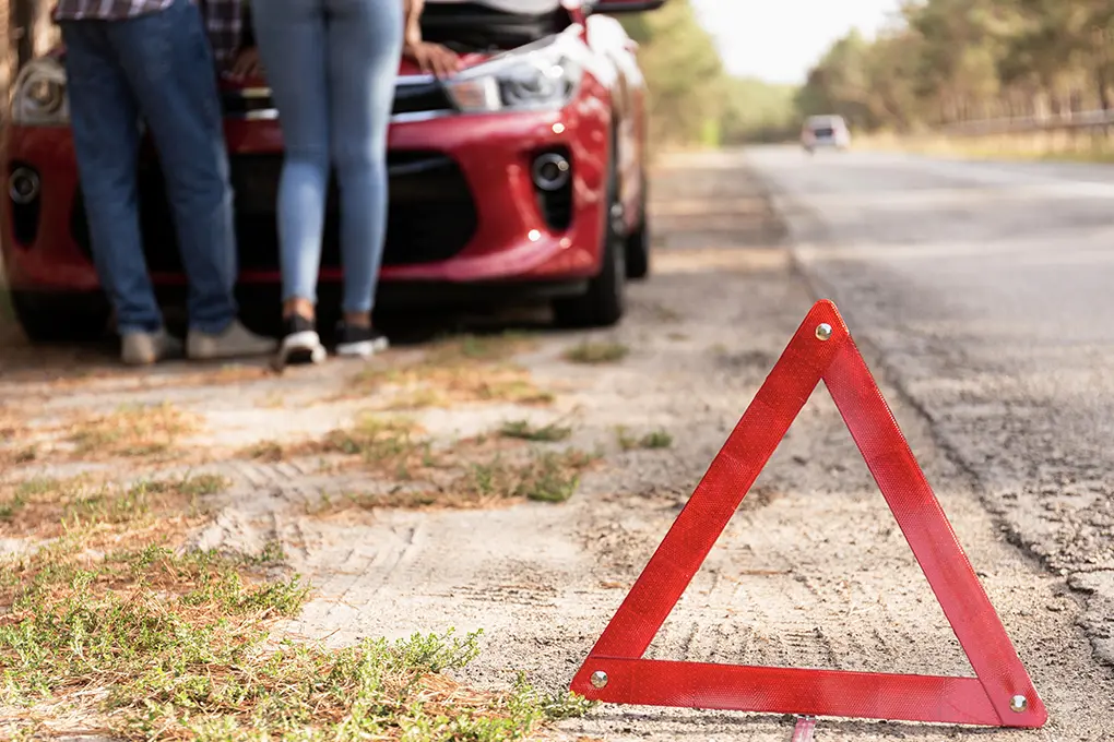 Red car with raised hood on roadside & a red warning triangle in foreground. Two people examine the engine, conveying concern and urgency.