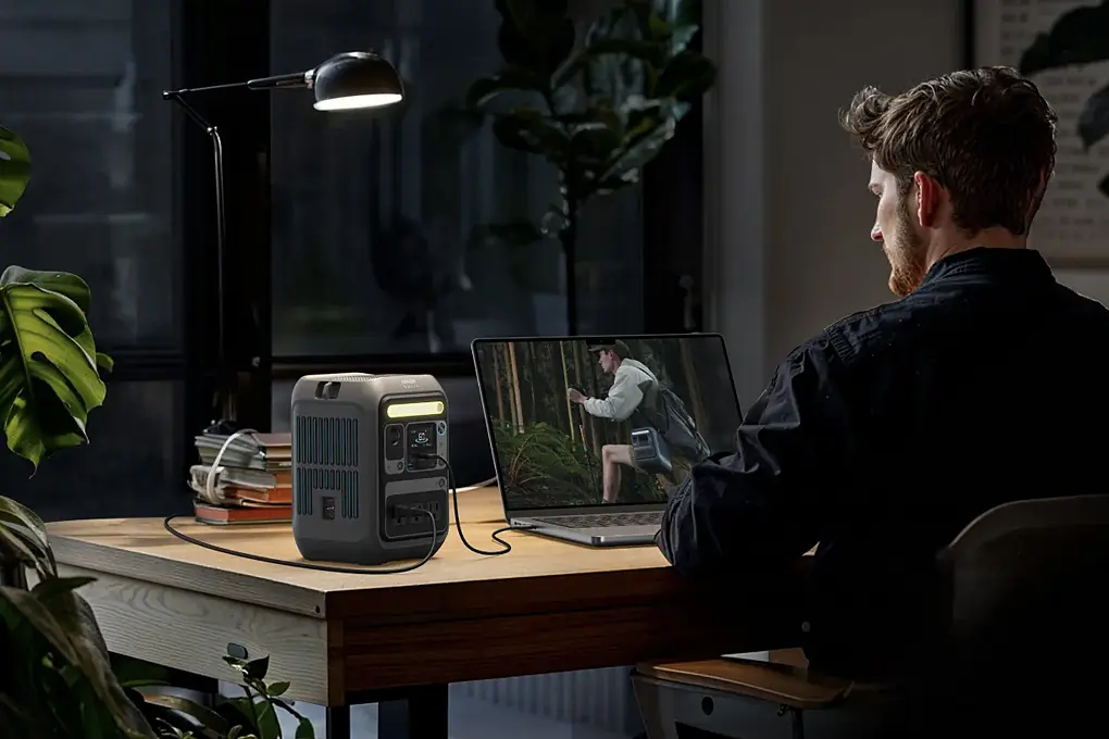 A man is using a laptop on a table with a table lamp, both connected to an Anker SOLIX C300X Portable Power Station